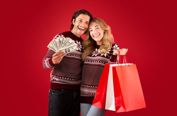 Compras de Navidad. Preciosa pareja joven en ropa de invierno sosteniendo el dinero y bolsas de regalo sobre fondo rojo - Foto, Imagen
