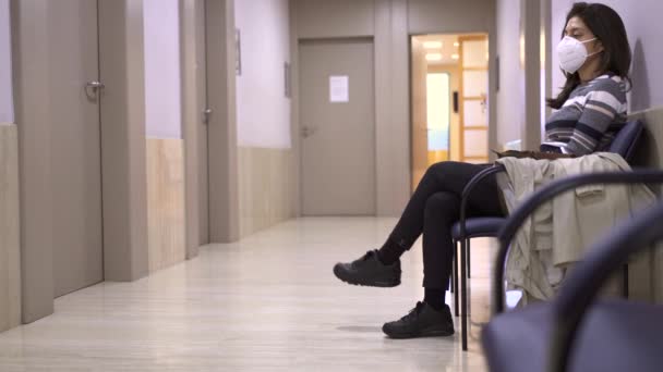 Paziente donna con maschera facciale seduta nella sala d'attesa vuota dell'ospedale. Giovane donna nervosa dal medico durante la pandemia di Coronavirus - Filmati, video