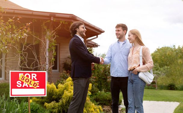 Cheerful realtor shaking hands with clients in front of residential building outdoors, free space - Photo, Image