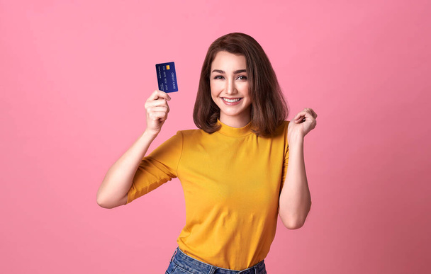 Junge Frau hält Kreditkarte in lässigem gelben Shrit mit wunderschönem Vertrauen und Zuversicht für Geldtransaktionen isoliert auf leuchtend rosa Hintergrund. - Foto, Bild