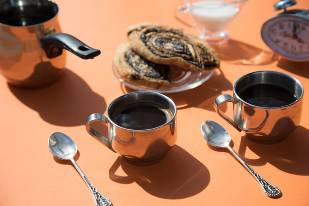 Sobre un fondo naranja en una mañana soleada, hay tazas de café de metal, una cafetera, rollos en un plato, un lechero y un despertador. Orientación horizontal - Foto, Imagen