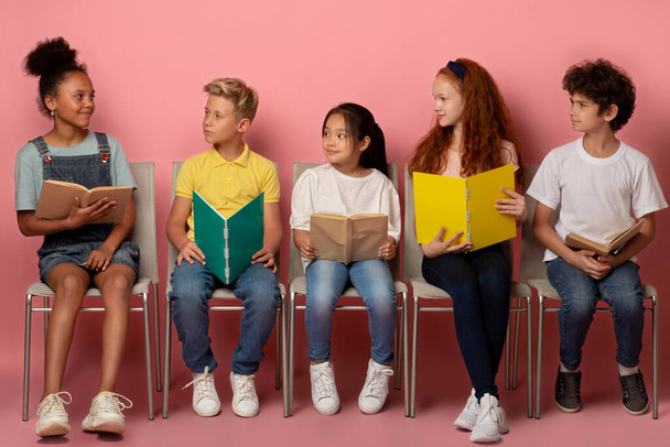 Przemysłowe wielonarodowe dzieci w wieku szkolnym z materiałami do nauki siedzącymi na krzesłach na różowym tle - Zdjęcie, obraz
