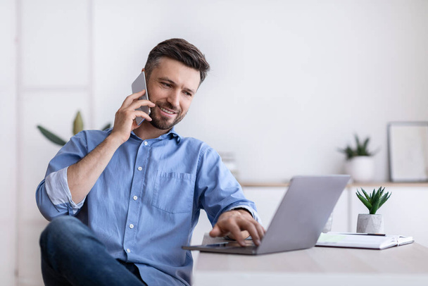 Веселий молодий чоловік працює дистанційно з домашнього офісу з мобільним телефоном та ноутбуком
 - Фото, зображення