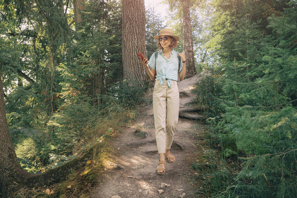 Счастливая азиатка с мобильным телефоном ходит по тропинке в лесу национального парка. Мобильное приложение с картами и доступом в Интернет в лесу - Фото, изображение