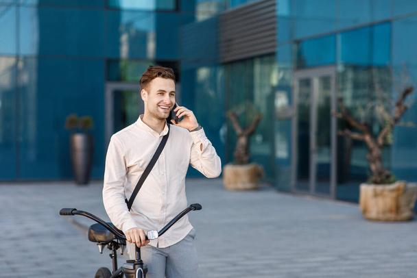 Conectado con amigos, al aire libre. Sonriente hombre habla por teléfono inteligente y pasea con la bicicleta - Foto, Imagen
