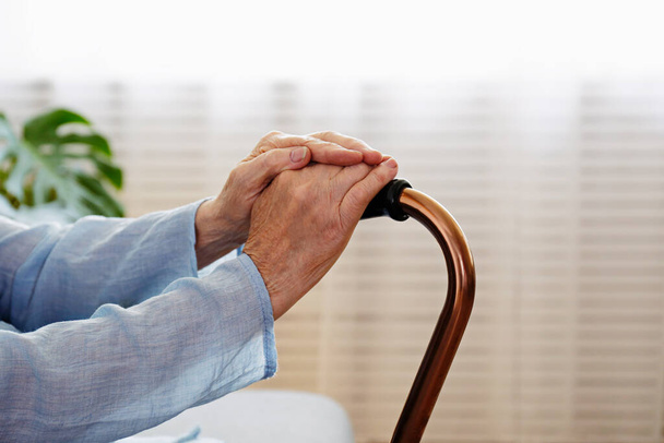Elderly woman sitting in nursing home room holding walking quad cane with wrinkled hand. Old lady wearing beige cardigan, metal aid stick handle bar close up. Interior background, copy space - Photo, Image