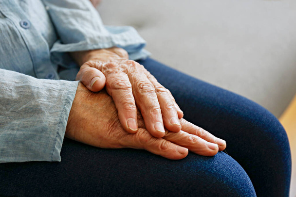 Elderly woman sitting alone at home, with hands folded on her laps. Senior old lady experiencing severe arthritis rheumatics pains, massaging, warming up arm. Close up, copy space, background - Photo, Image