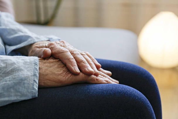 Elderly woman sitting alone at home, with hands folded on her laps. Senior old lady experiencing severe arthritis rheumatics pains, massaging, warming up arm. Close up, copy space, background - Foto, immagini