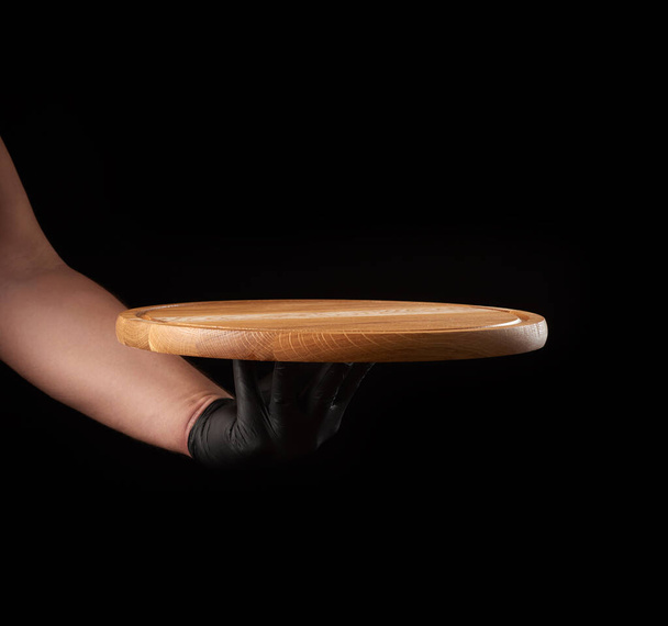 мужская рука в черной латексной перчатке держит круглый пустой деревянной доски пиццы, черный фон - Фото, изображение