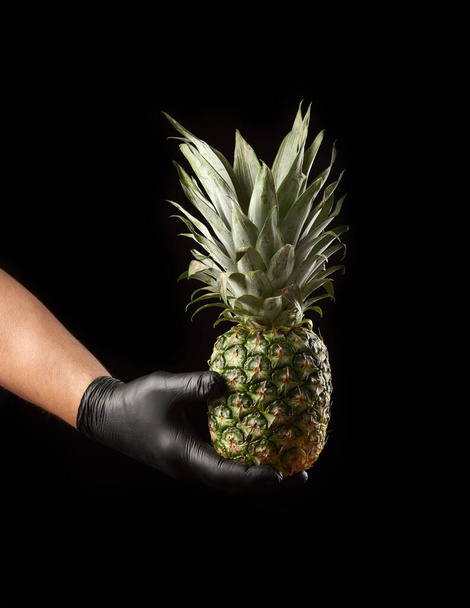 male hand in black latex glove holds a whole unpeeled pineapple, black background - Photo, Image