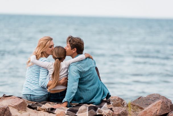 Back view of parents embracing and kissing daughter on plaid near sea  - Valokuva, kuva