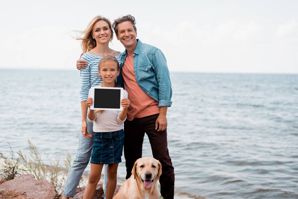 Girl holding digital tablet near parents and golden retriever on seaside - Foto, Imagem