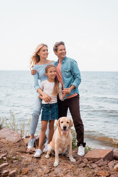 Selective focus of man embracing wife and daughter near golden retriever on seaside  - Foto, Imagem