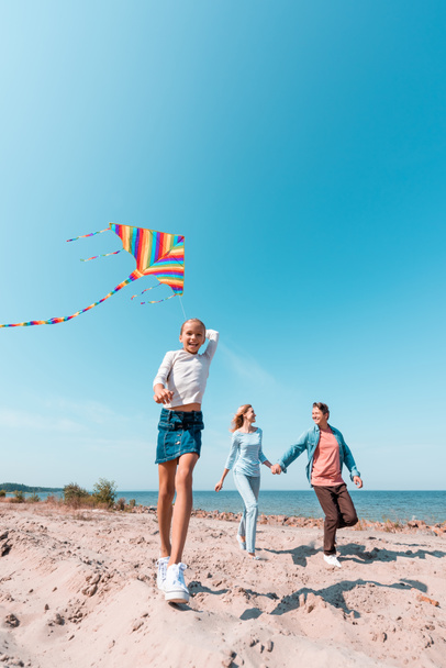 Selectieve focus van meisje houden vlieger in de buurt van ouders op het strand  - Foto, afbeelding