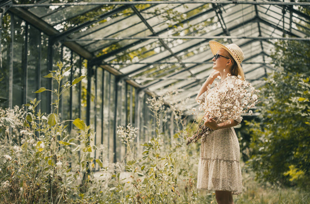 woman with a bouquet of dried flowers in a retro dress and a hat in a old greenhouse - Foto, afbeelding