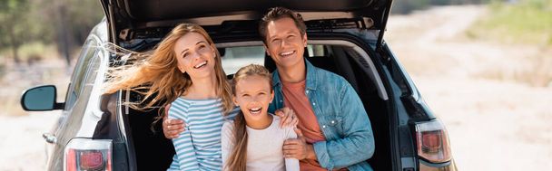 Панорамный снимок семьи, смотрящей в камеру, сидящей в багажнике автомобиля на улице  - Фото, изображение