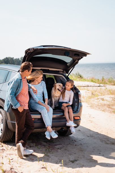 Selective focus of parents looking at daughter with golden retriever in car trunk on beach  - Φωτογραφία, εικόνα