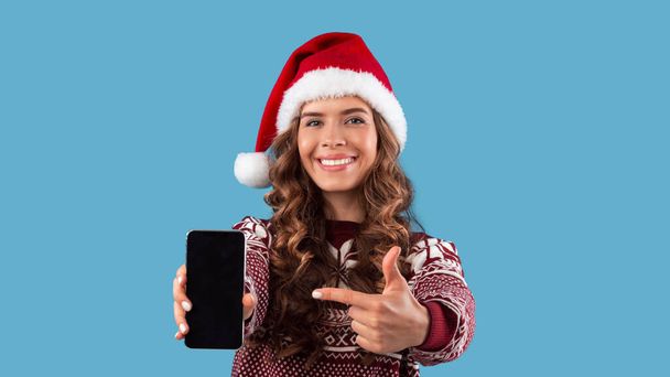 Ελκυστική νεαρή κοπέλα με στολή Χριστουγέννων που δείχνει το smartphone με λευκή οθόνη σε μπλε φόντο, mockup για το σχεδιασμό - Φωτογραφία, εικόνα