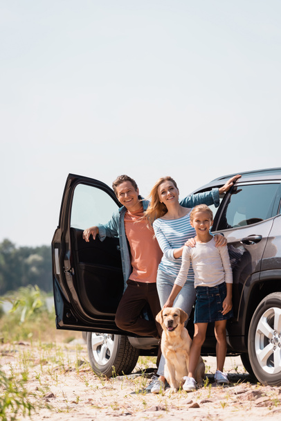 Selective focus of family with golden retriever looking at camera while standing near car outdoors  - Photo, Image