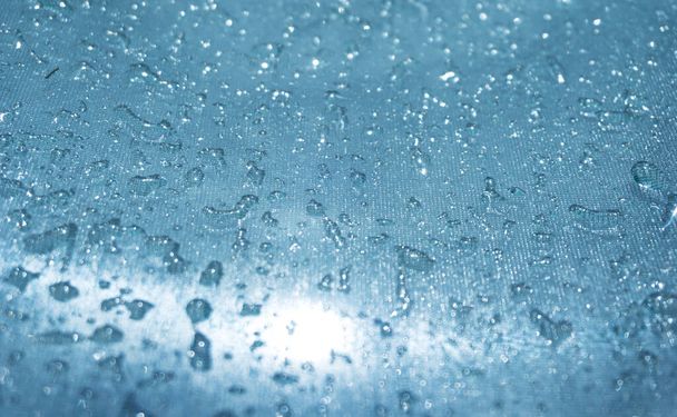 Σταγόνες βροχής στο μπλε γυάλινο φόντο bokeh, γυαλιστερές σταγόνες βροχής σε μια γυάλινη επιφάνεια, σταγόνες νερού σε μια πισίνα closeup - Φωτογραφία, εικόνα
