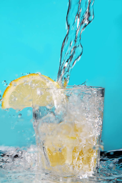 Water pouring into the glass with lemon over blue background - Photo, Image