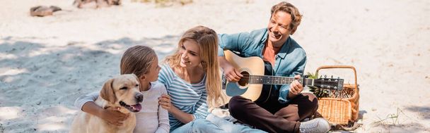 Panoramic shot of woman embracing child near golden retriever and husband with acoustic guitar on beach  - Photo, Image