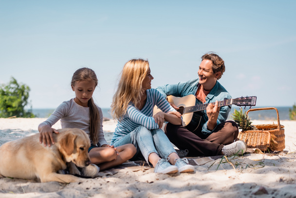 Selektiver Fokus der Frau auf Ehemann mit Akustikgitarre neben Tochter mit Golden Retriever am Strand  - Foto, Bild