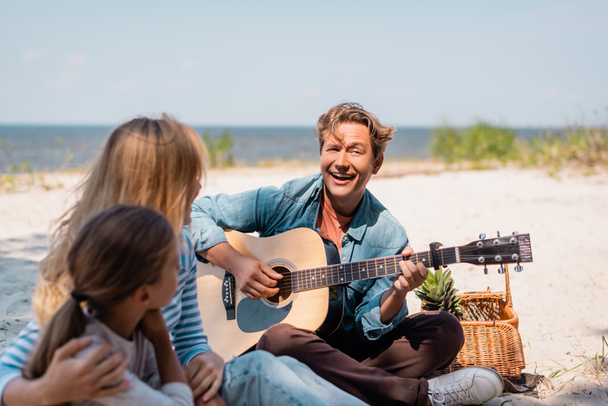Selective focus of man playing acoustic guitar near wife and daughter on beach  - Фото, изображение