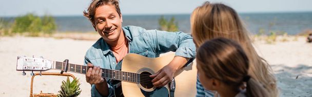 Horizontal image of man playing acoustic guitar near family on beach  - Photo, Image
