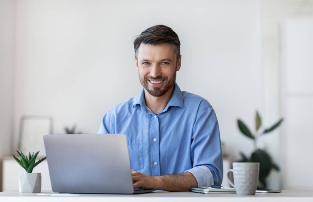 Jeune homme d'affaires souriant assis au bureau avec ordinateur portable dans le bureau moderne - Photo, image