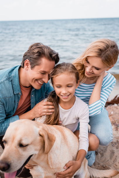 Selective focus of father hugging daughter near wife and golden retriever on beach  - Foto, Imagen