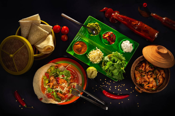 Spicy chicken, vegetables, sour cream, cheese, guacamole, Enchilada Sauce and corn tortillas fajitas in bamboo pancake basket on a dark background, top view. Delicious snack, tapas in mexican style - Photo, Image