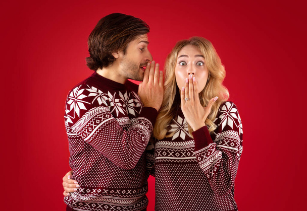 Shocked young lady in Christmas sweater and boyfriend whispering secret into her ear on red background - Photo, Image