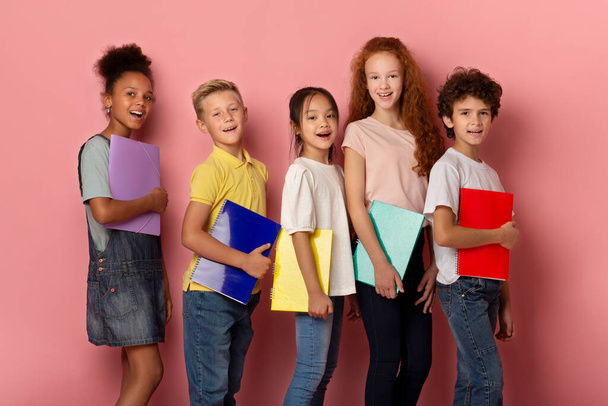Vreugdevolle jongens en meisjes van verschillende nationaliteiten met copybooks poseren over roze achtergrond - Foto, afbeelding