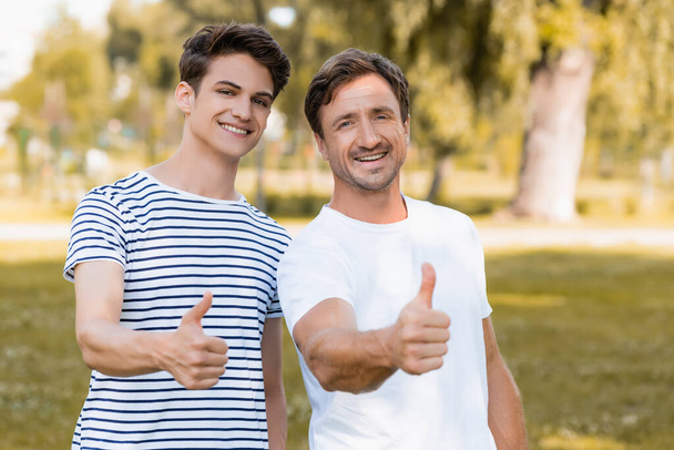 joyful father and teenager son in t-shirts showing thumbs up in park - Fotografie, Obrázek