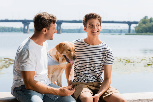 father looking at pleased son near golden retriever and river - Foto, Imagem