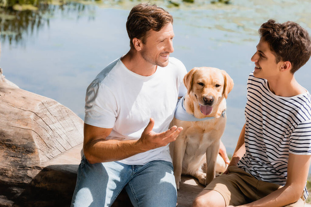 father and son looking at each other while sitting near golden retriever and lake - Photo, Image