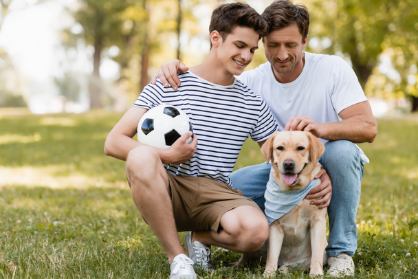 father sitting near teenager son with football cuddling golden retriever - Foto, imagen