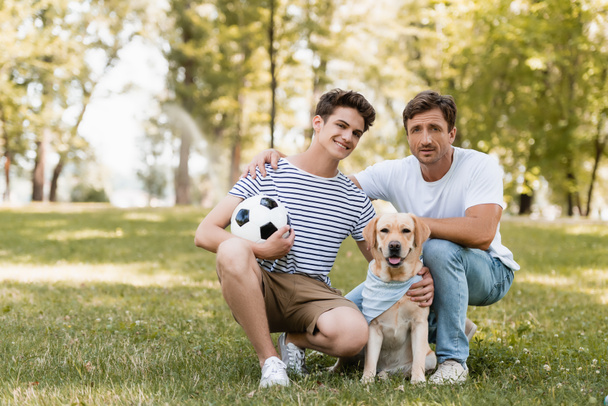 otec sedí poblíž potěšený teenager syn s fotbalem a zlatý retrívr - Fotografie, Obrázek