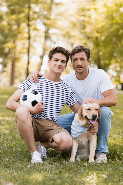 father sitting on grass near teenager son with football and golden retriever - Φωτογραφία, εικόνα