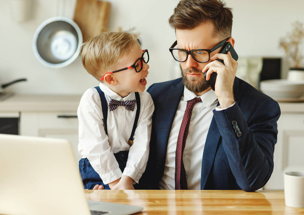 Knappe jonge zakenman in formele kleding en bril zitten aan tafel met vrolijke kleine zoon op de knieën en praten op de smartphone tijdens het werken online bij hom - Foto, afbeelding