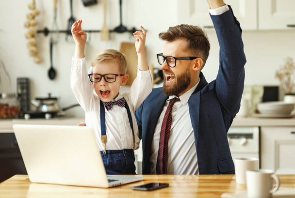 Молодой предприниматель в формальной одежде и очках с маленьким сыном в формальной одежде смотрит на экран ноутбука и празднует успешную сделку, работая удаленно в современных квартирах - Фото, изображение