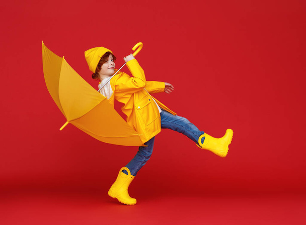 Side view of funny boy in raincoat and boots carrying umbrella on shoulder while walking against red backdrop on rainy da - Foto, Imagen