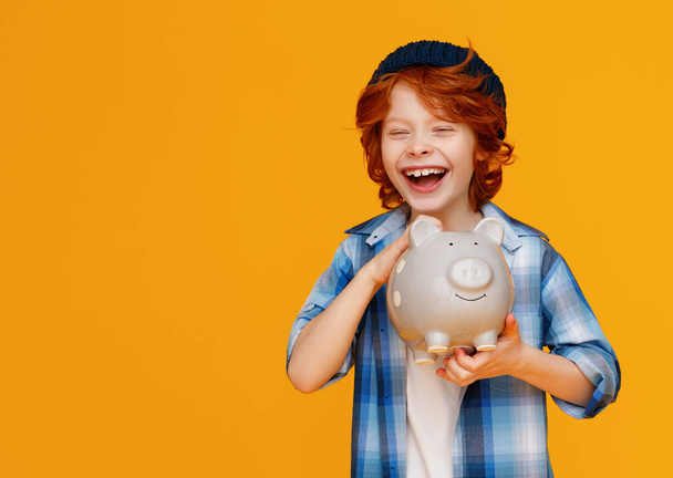 pozytywny imbir chłopiec w dorywczo strój śmiejąc się i oszczędzając pieniądze w świnka bank na żółtym tle - Zdjęcie, obraz
