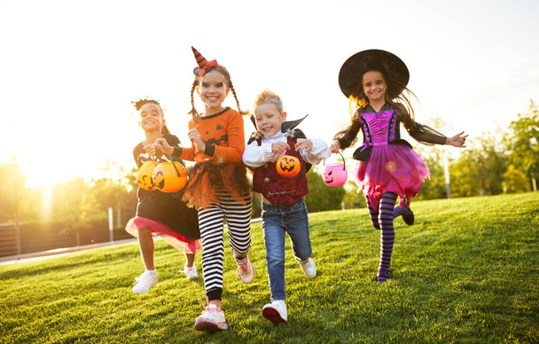 Grupo de crianças animadas em fantasias assustadoras sorrindo e correndo no gramado durante a celebração de Halloween à noite, a par - Foto, Imagem