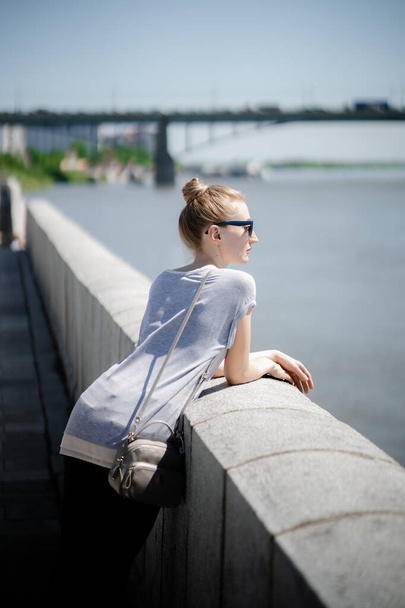Egy napszemüveges fiatal nő áll a folyó partján egy híd hátterében egy napos nyári napon. Önálló gondolatok, nyaralás városi környezetben vagy nyári séta koncepció - Fotó, kép