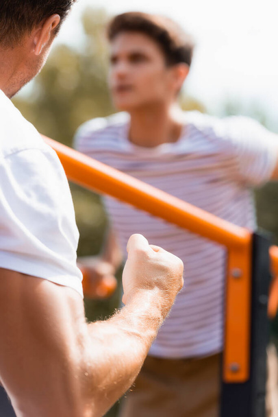selective focus of father with clenched fist near teenager son exercising on horizontal bars  - Foto, Imagen