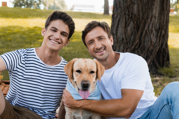 joyful father and teenager son looking at camera near golden retriever - Photo, Image