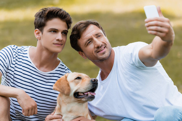 father and teenager boy taking selfie with golden retriever in park - Valokuva, kuva