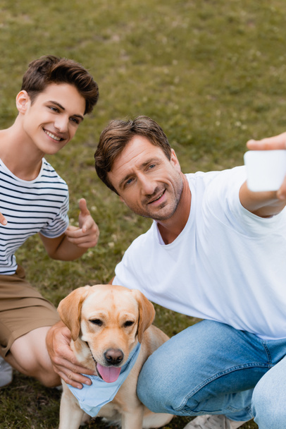 selective focus of teenager boy pointing with finger at golden retriever while father taking selfie - Fotografie, Obrázek